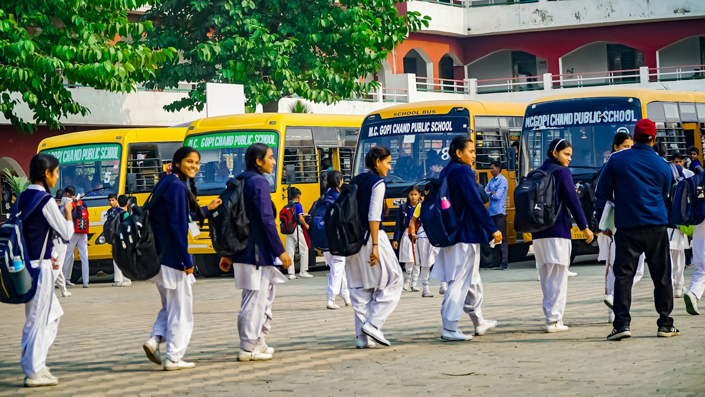Students in uniforms walk past yellow school buses.